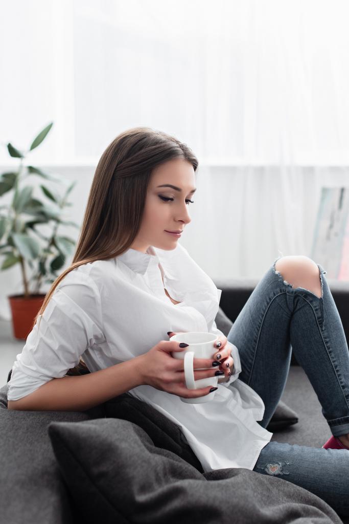 chica pensativa sentada en el sofá y sosteniendo la taza de café en la sala de estar
 - Foto, Imagen