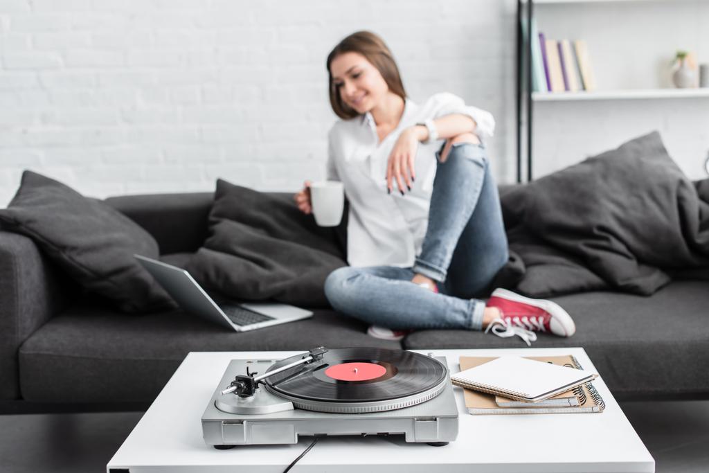 meisje in wit overhemd zittend op de Bank met een koffiekopje, met behulp van laptop en vinyl platenspeler in de woonkamer beluisteren - Foto, afbeelding