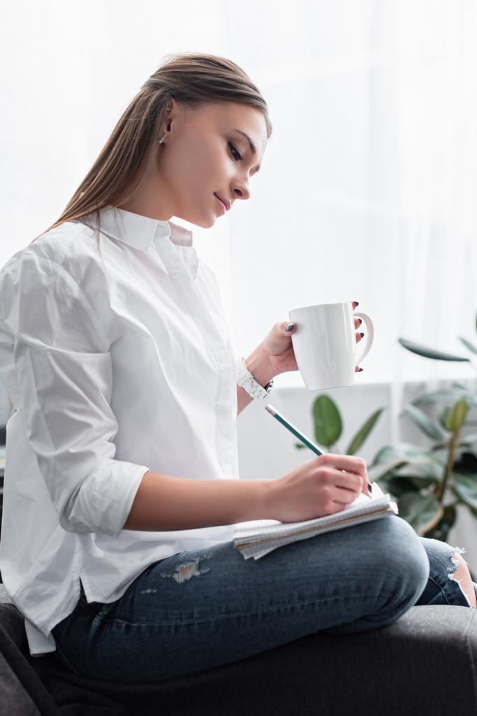 attraente ragazza in camicia bianca seduta con tazza di caffè e scrittura in notebook a casa
 - Foto, immagini
