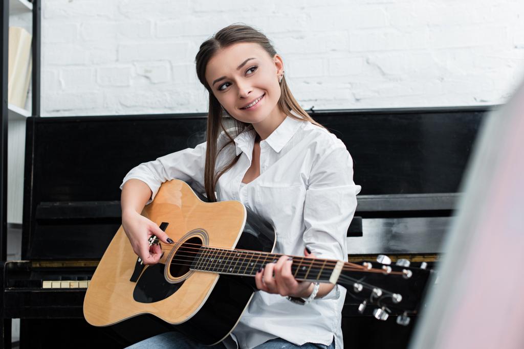 glimlachend meisje zit en speel gitaar in woonkamer met piano op de achtergrond - Foto, afbeelding