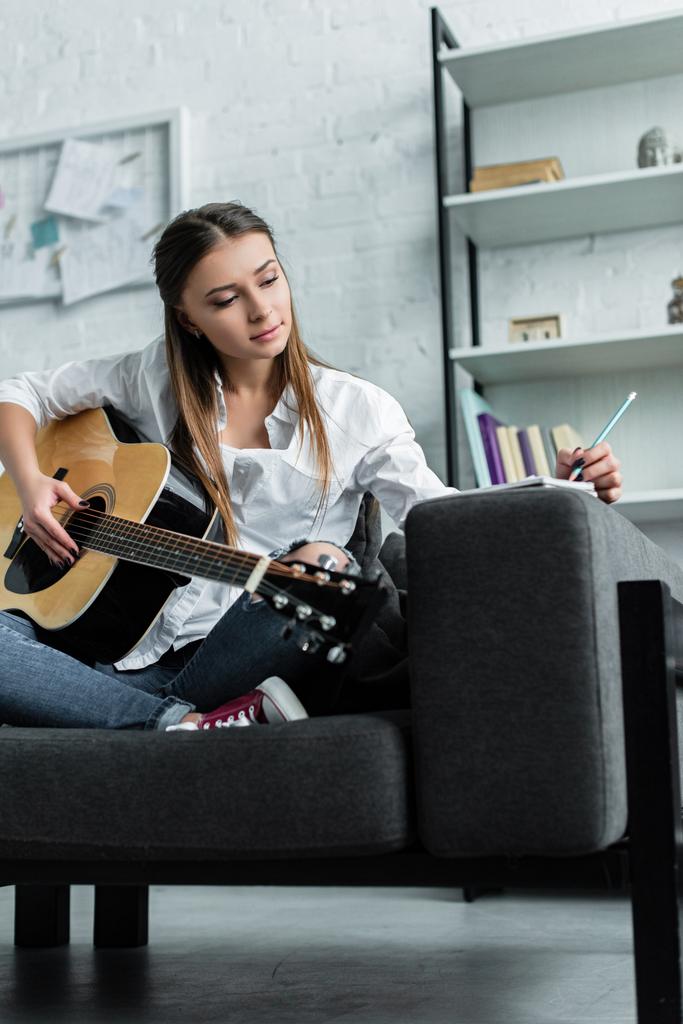 pensive girl sitting on couch with guitar, writing in notebook and composing music in living room - Photo, Image