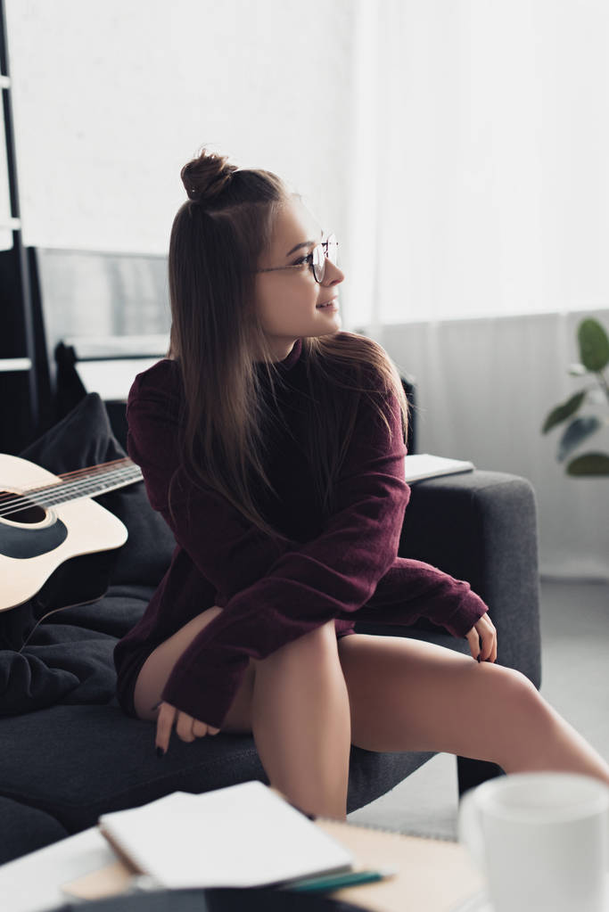beautiful smiling girl in glasses looking away and sitting on couch in living room - Photo, Image