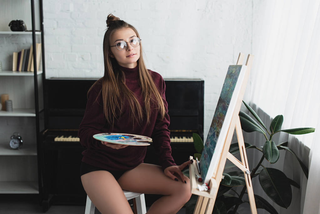 Mädchen im weinroten Pullover sitzt und schaut in die Kamera, während sie zu Hause malt  - Foto, Bild