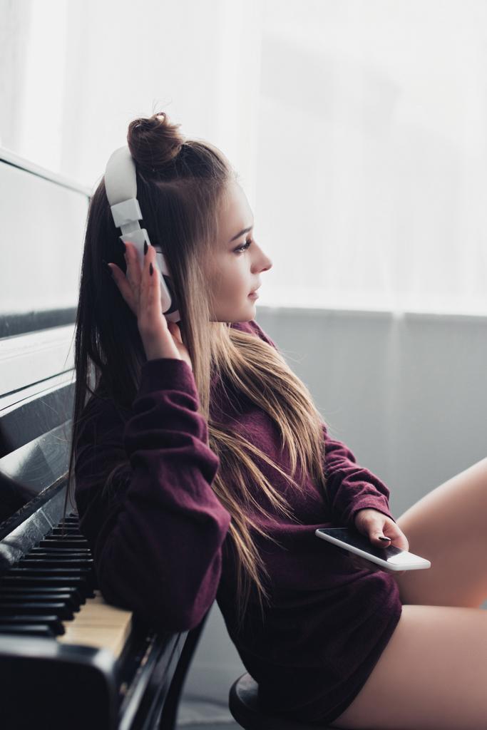 attractive girl with headphones on head sitting on chair in front of piano and holding smartphone at home - Photo, Image