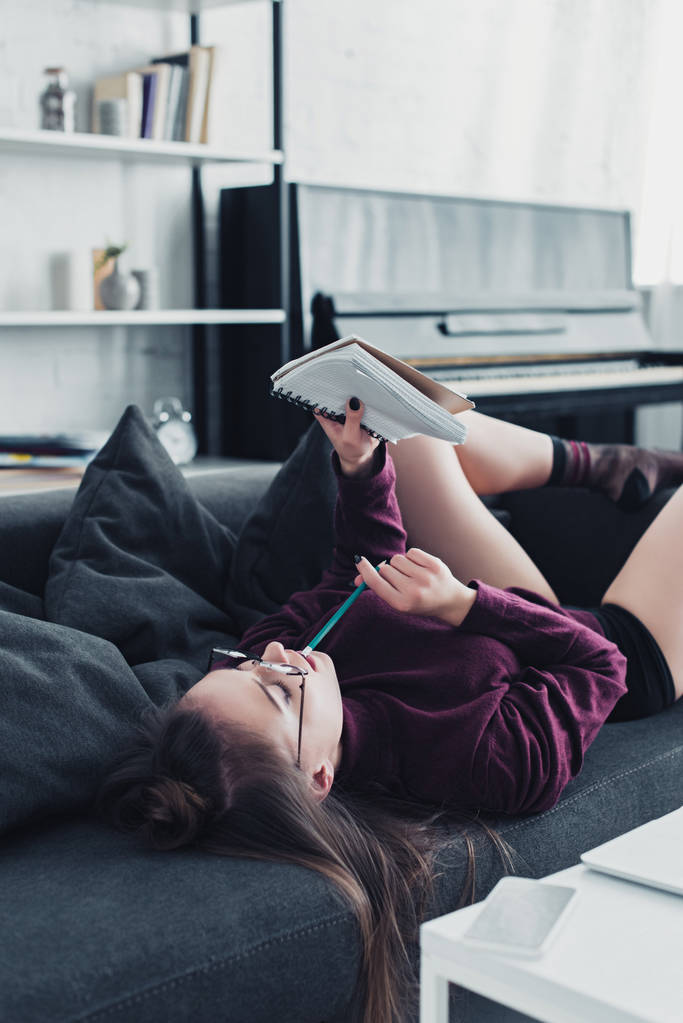 beautiful girl in glasses lying on sofa, holding pencil and looking at notebook in living room - Photo, Image