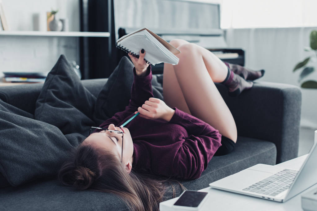 attractive girl lying on sofa, holding pencil and looking at notebook in living room - Photo, Image