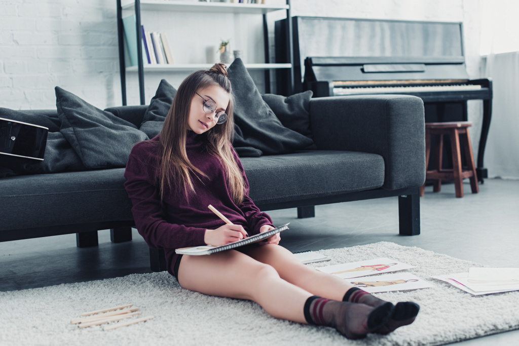 gyönyörű tervező ül a szőnyegen közelében a kanapén, és ceruzával a notebook otthon - Fotó, kép