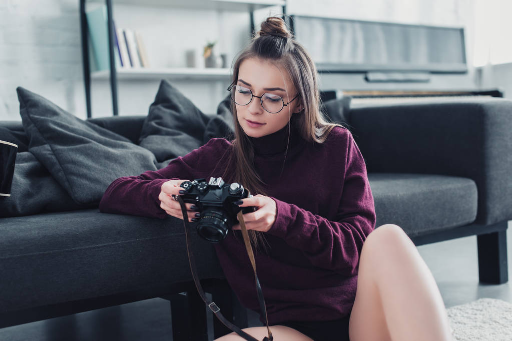 attractive photographer sitting on floor near couch and holding film camera in living room - Photo, Image