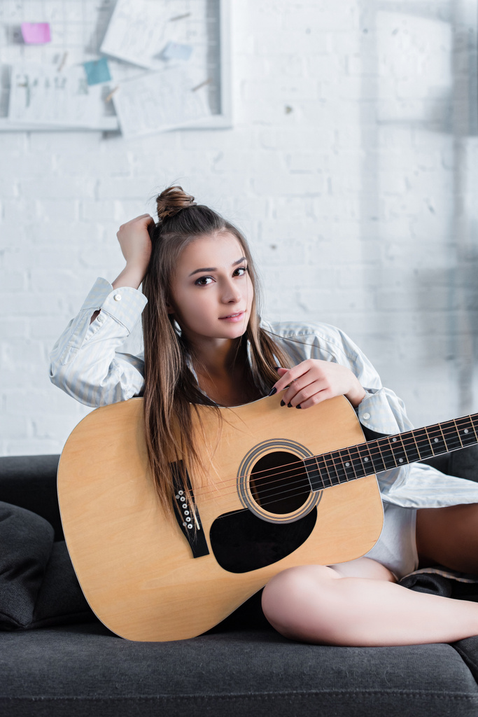 attractive musician sitting on sofa, holding acoustic guitar and looking at camera at home - Photo, Image