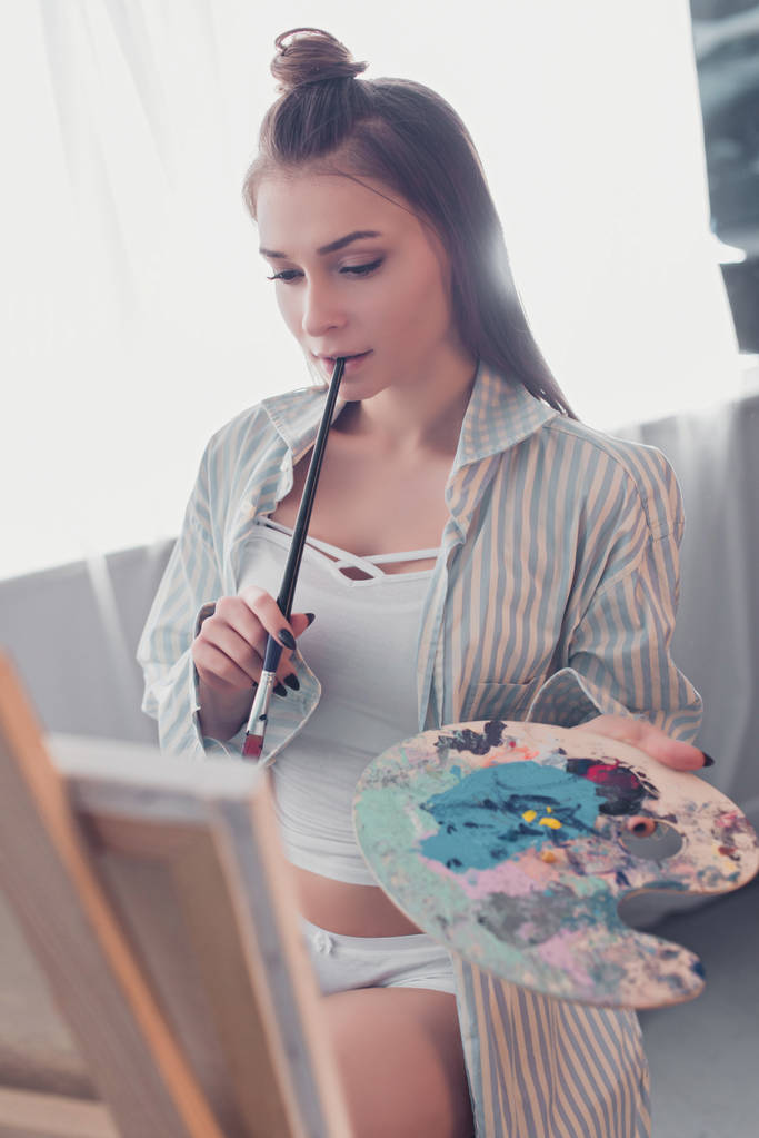 attrayant artiste tenant palette, pinceau mordant et regardant chevalet dans le salon
 - Photo, image