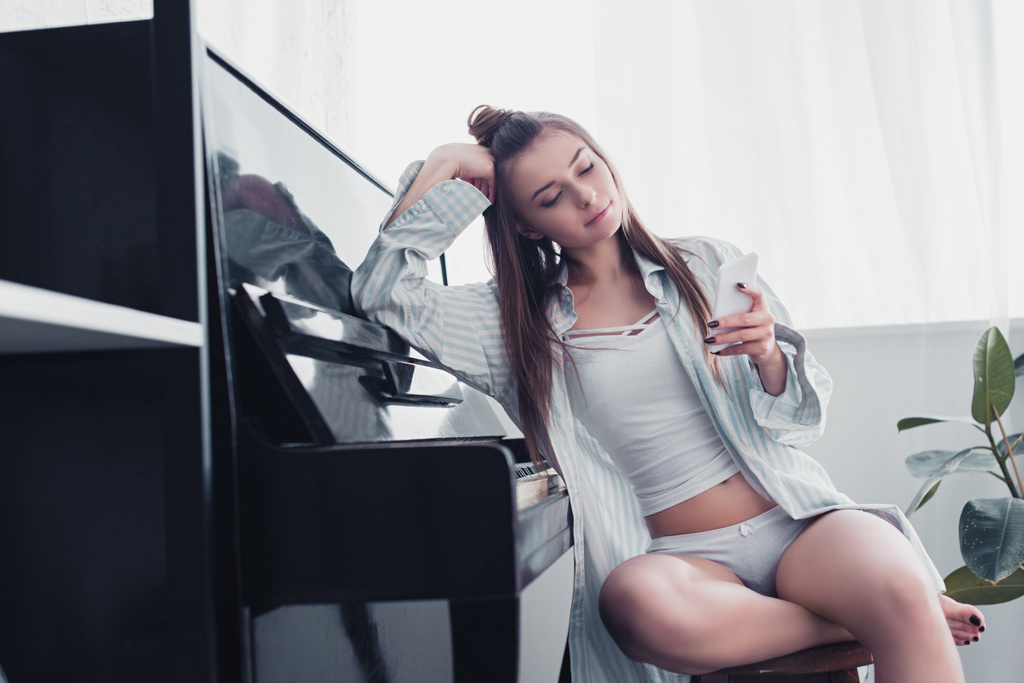 sexy Mädchen in Hemd und Unterwäsche sitzt am Klavier und benutzt Smartphone zu Hause - Foto, Bild