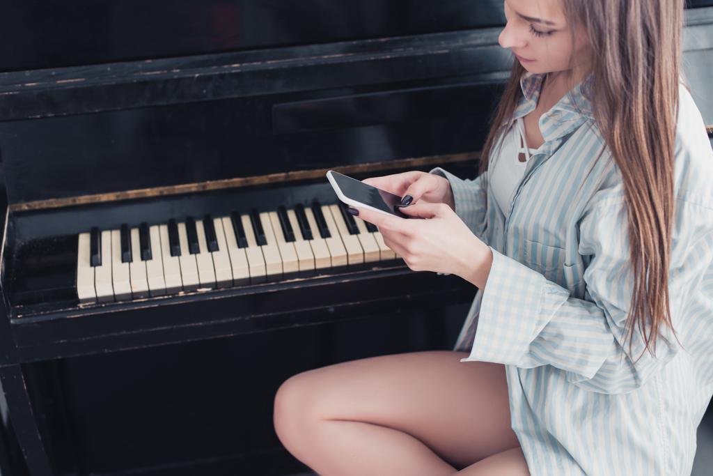 attraktives Mädchen im Hemd sitzt vor dem Klavier und benutzt Smartphone im Wohnzimmer - Foto, Bild