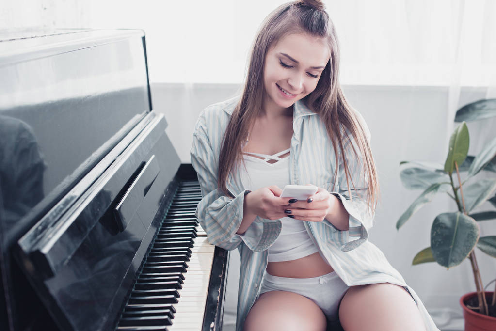 menina bonita em roupa interior e camisa sentada na frente do piano e usando smartphone na sala de estar
 - Foto, Imagem