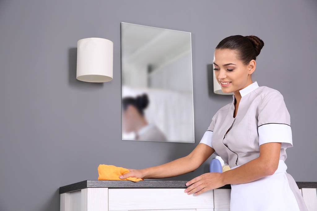 Jovem camareira limpando poeira de móveis com pano no banheiro
 - Foto, Imagem