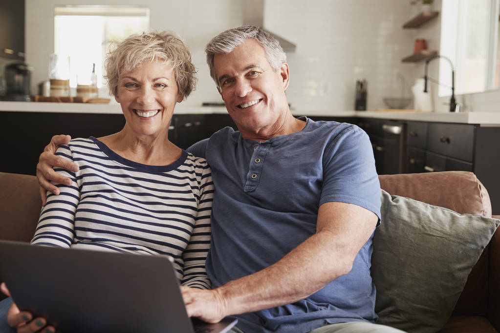 Senior paar zittend op Bank laptop, lacht om de camera gebruiken - Foto, afbeelding
