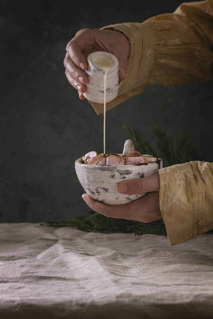 Keramische Tasse heiße Schokolade mit Marshmallow s 'mores in weiblichen Händen. Sahne aus Krug gießen. graue Tischdecke aus Leinen. Winterdrink. Düstere Atmosphäre. - Foto, Bild