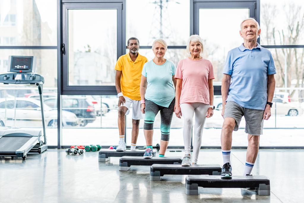 Glückliche multiethnische Senioren stehen in der Nähe von Trittbrettern im Fitnessstudio - Foto, Bild
