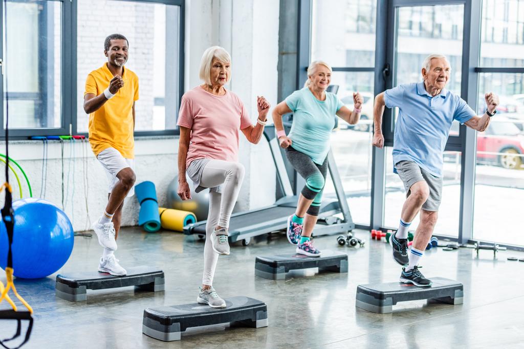 fröhliche multiethnische Senioren beim Synchrontraining auf Trittbrettern im Fitnessstudio - Foto, Bild