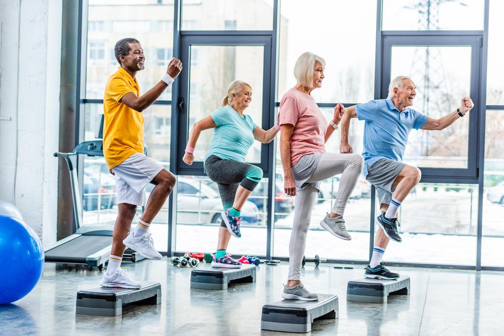 Seitenansicht multiethnischer Senioren-Athleten beim Synchrontraining auf Trittplattformen im Fitnessstudio - Foto, Bild