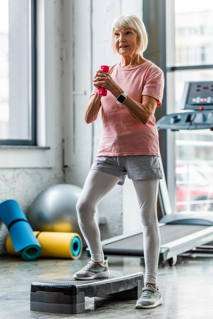 senior sportswoman with smartwatch making exercise with dumbbell on step platform at gym - Photo, Image