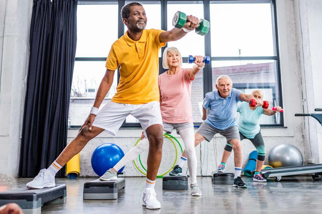 selective focus of senior multicultural sportspeople synchronous exercising with dumbbells on step platforms at gym - Photo, Image