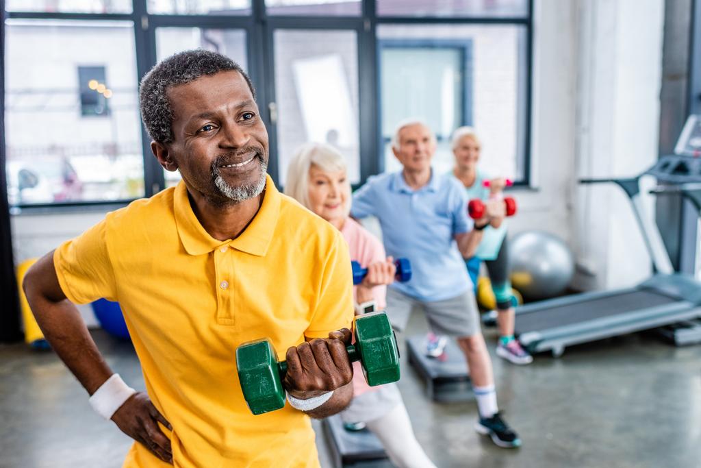african american man and his friends synchronous exercising with dumbbells at sports hall - Photo, Image
