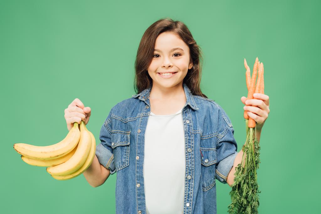 child looking at camera and holding bananas with carrots isolated on green - Photo, Image