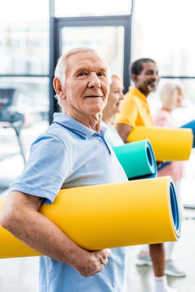 selective focus of senior sportsman holding fitness mat and his friends standing behind at gym - Photo, Image