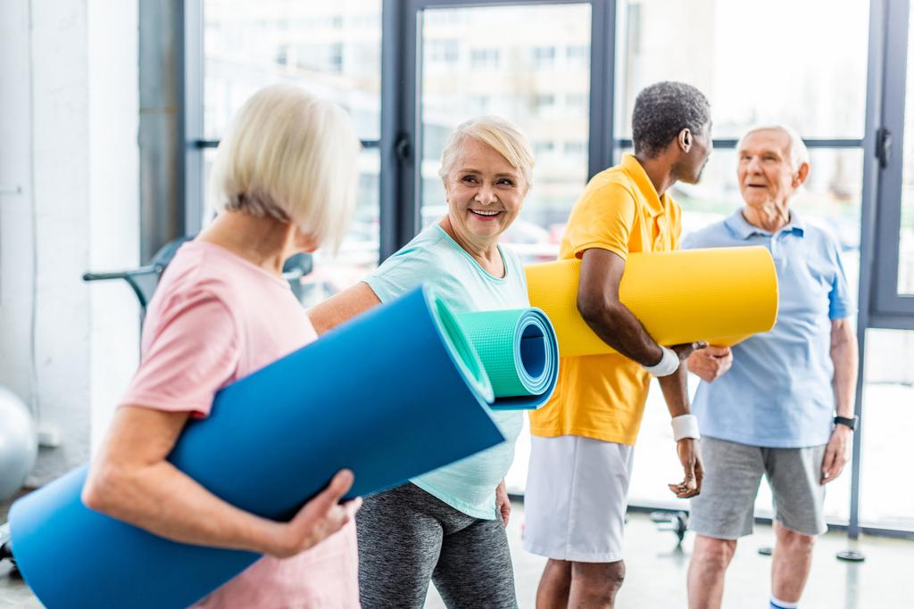senior sportswomen holding fitness mats and their male friends standing behind at gym - Photo, Image