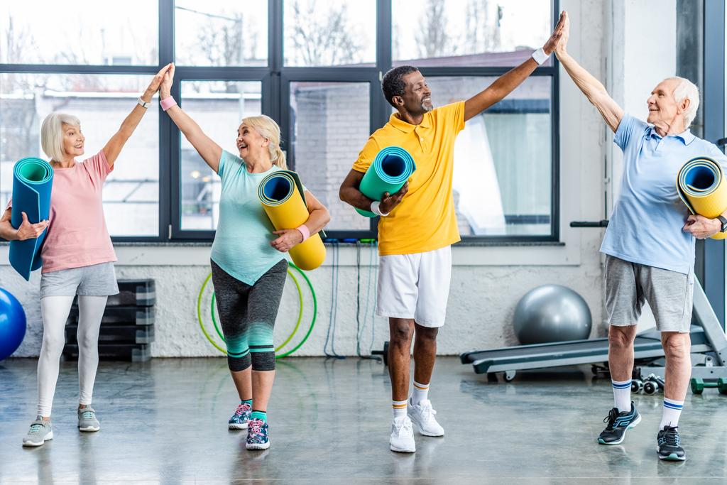 смеющиеся старшие мультиэтнические спортсмены с фитнес-ковриками, дающие друг другу пять в спортзале
 - Фото, изображение