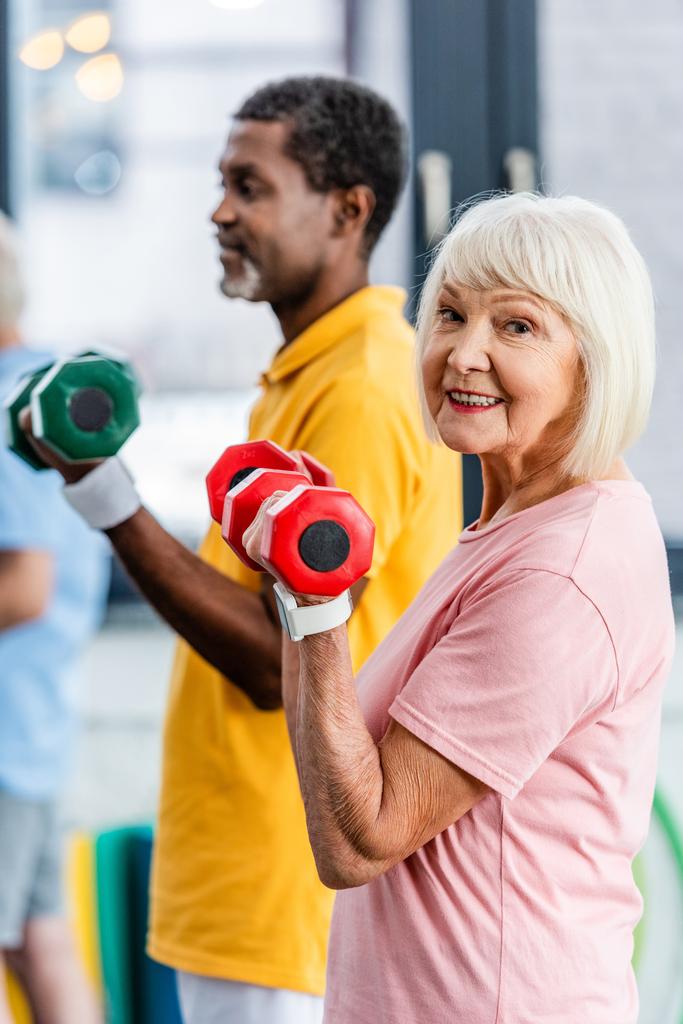 selective focus of senior woman and her african american husband doing exercise with dumbbells at gym - Photo, Image