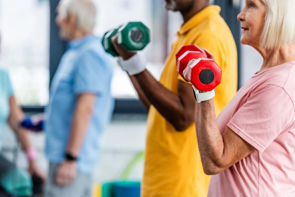 vista parcial de pareja madura interracial haciendo ejercicio con pesas en el gimnasio
 - Foto, Imagen