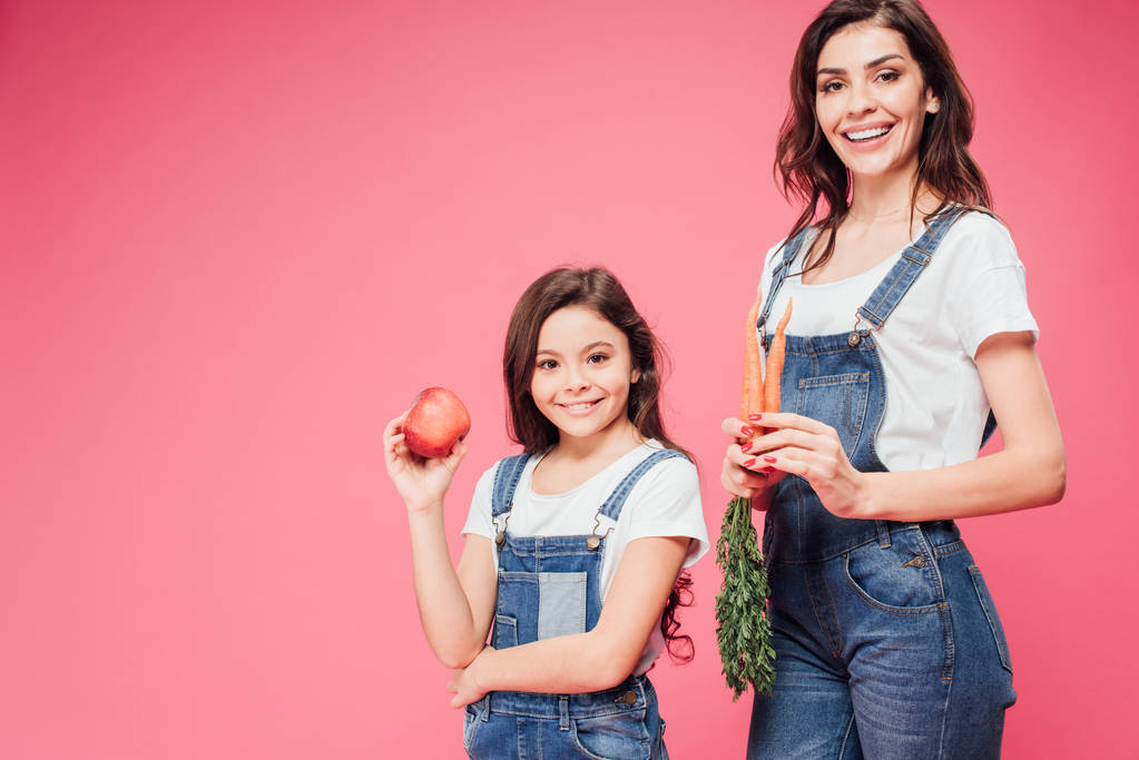 glückliche Mutter und Tochter halten roten Apfel und Karotten isoliert auf rosa  - Foto, Bild