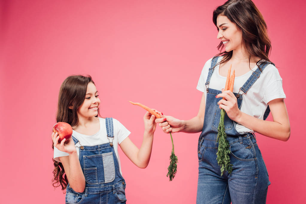 vrolijke moeder wortel geven aan dochter geïsoleerd op roze  - Foto, afbeelding