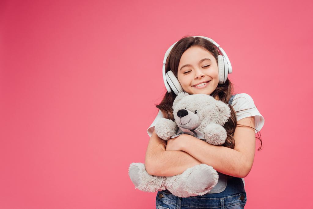 szczęśliwe dziecko w słuchawki przytulanie pluszowego misia na różowym tle  - Zdjęcie, obraz
