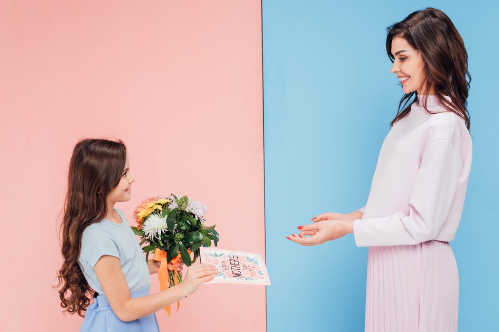 adorable child giving bouquet and greeting card to attractive woman on blue and pink background - Photo, Image