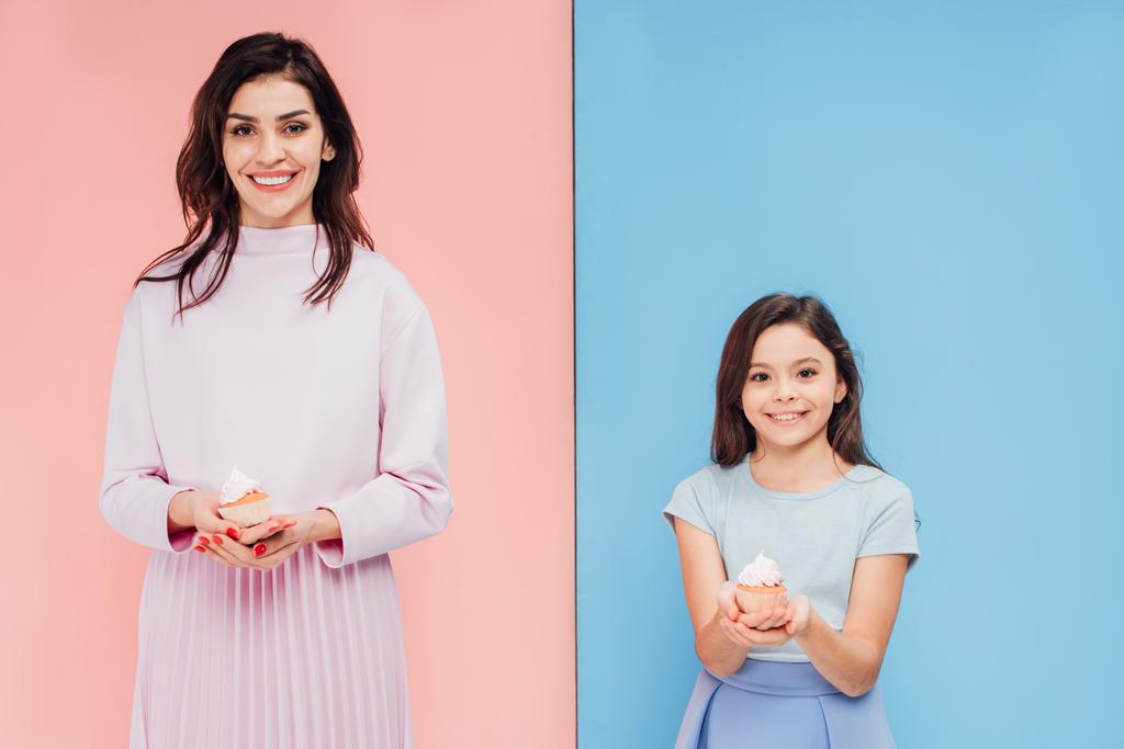 beautiful woman and child holding cupcakes and looking at camera on blue and pink background - Photo, Image