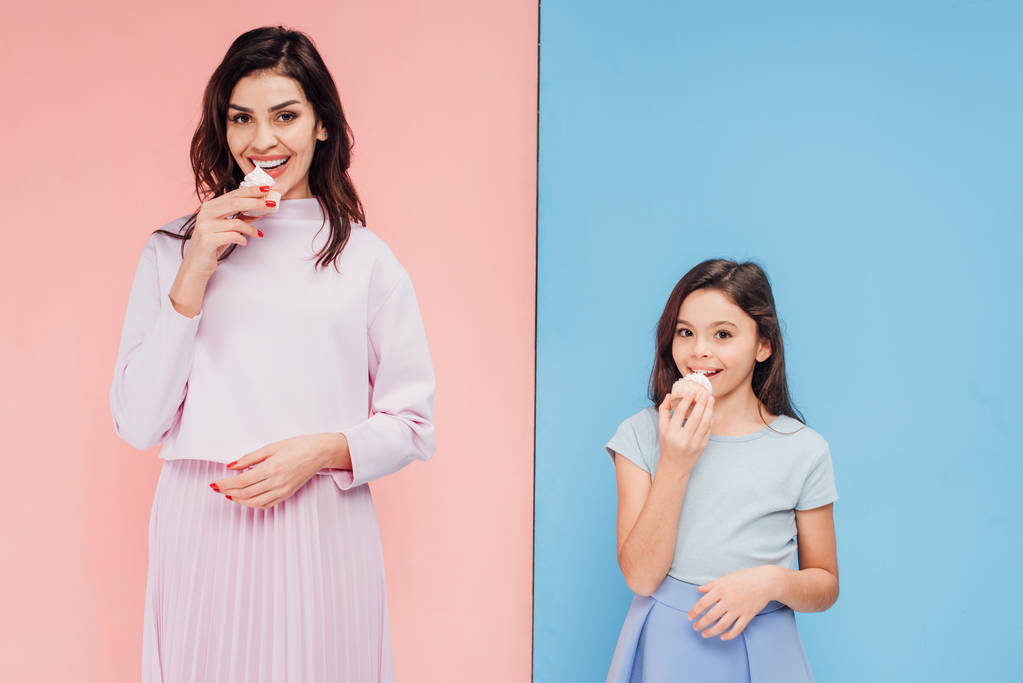 bela mulher e criança comendo cupcakes e olhando para a câmera em fundo azul e rosa
 - Foto, Imagem