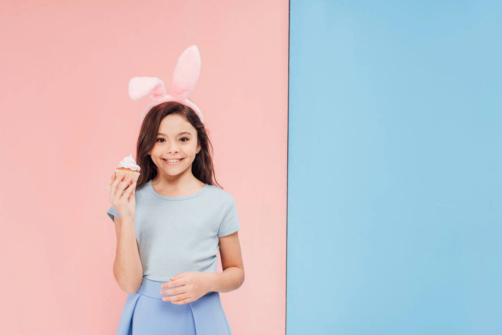 adorable enfant dans les oreilles de lapin de Pâques tenant cupcake et regardant la caméra sur fond bleu et rose
 - Photo, image