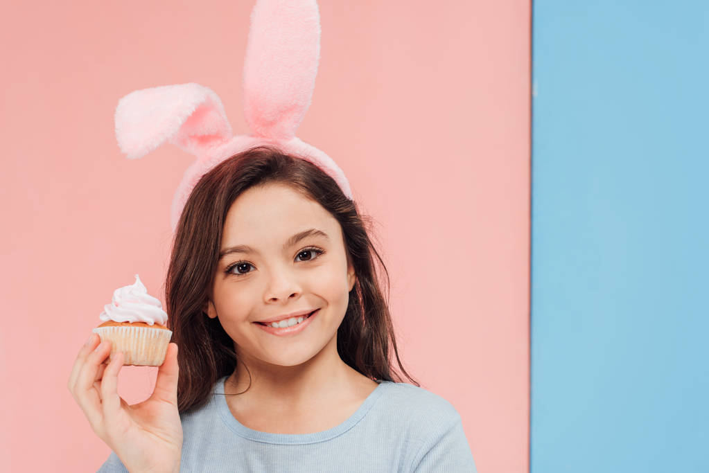 очаровательный ребенок в кроличьи уши, держа кекс и глядя на камеру на синем и розовом фоне
 - Фото, изображение