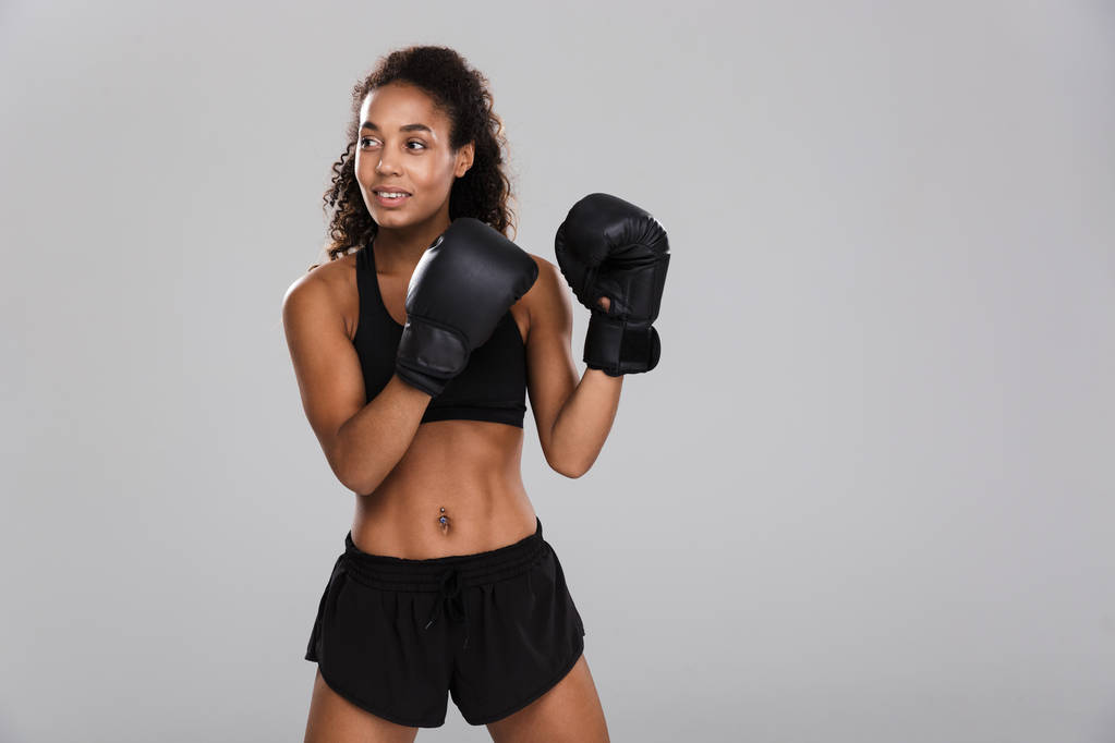 Портрет афро-американской молодой спортсменки, занимающейся боксом на сером фоне, отводящей взгляд
 - Фото, изображение