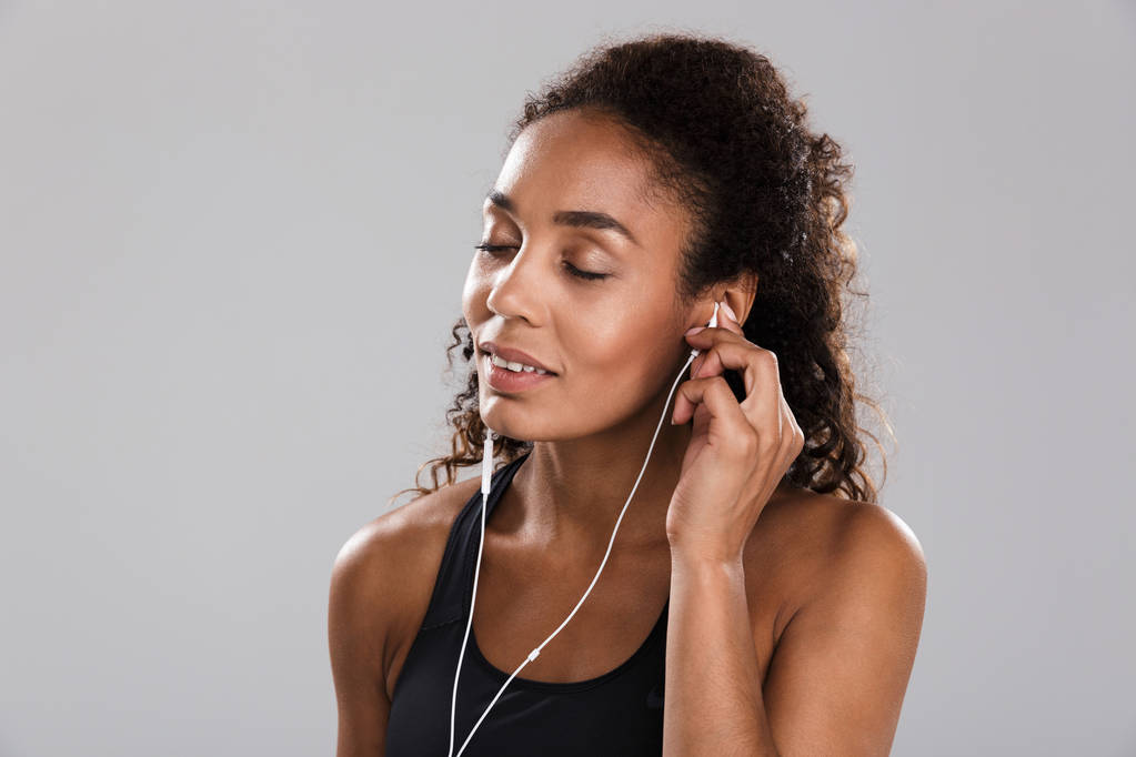 Portrait of an afro american young sportswoman isolated over gray background, listening to music with earphones - Photo, Image