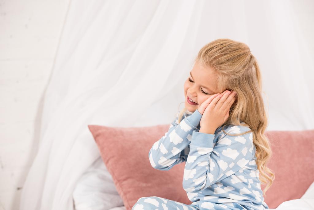 Niedliche schläfrige Kind sitzt auf dem Bett mit geschlossenen Augen und Händen in der Nähe der Ohren - Foto, Bild