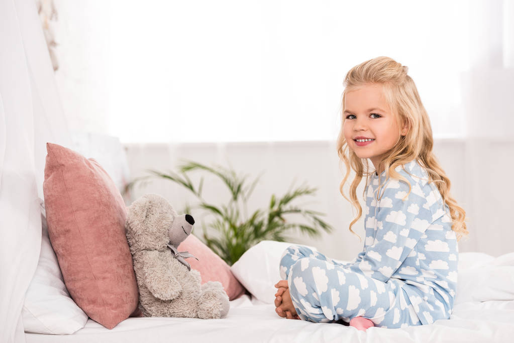 cute child sitting on bed with crossed legs and teddy bear - Photo, Image