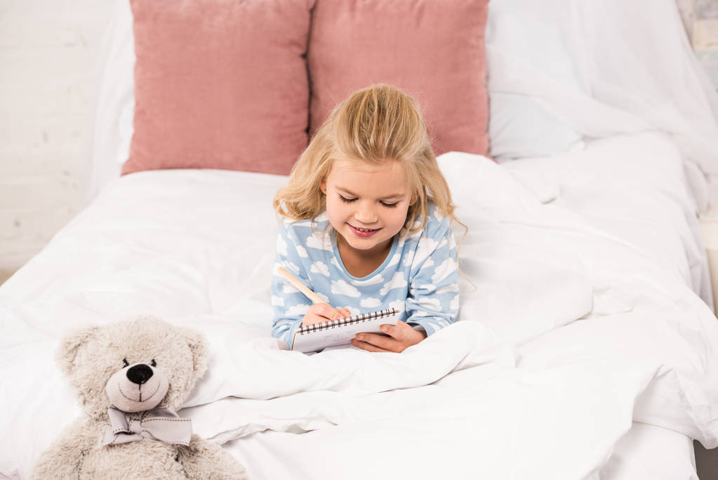 entzückendes Kind liegt im Bett und schreibt in Notizbuch - Foto, Bild