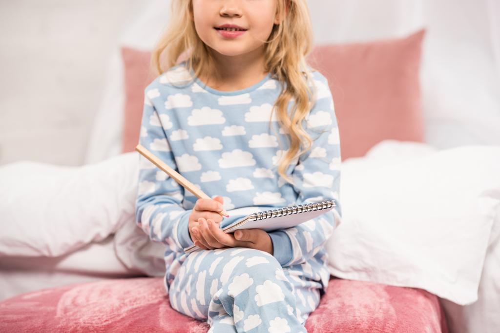 περικοπεί θέα του παιδιού στο πιτζάμες κάθεται στο κρεβάτι και γράφοντας στο Σημειωματάριο - Φωτογραφία, εικόνα
