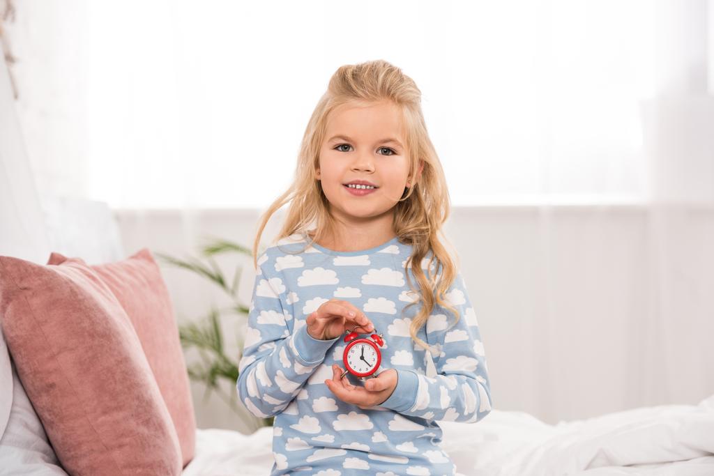 szczęśliwy adorable kid, siedząc na łóżku z małym zegarem w ręce - Zdjęcie, obraz