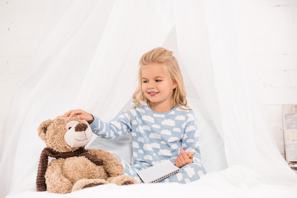 χαριτωμένο παιδί στο πιτζάμες κάθεται στο κρεβάτι με το σημειωματάριο και αρκουδάκι - Φωτογραφία, εικόνα