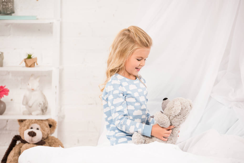 χαρούμενο παιδί παίζει με αρκουδάκι στο κρεβάτι - Φωτογραφία, εικόνα