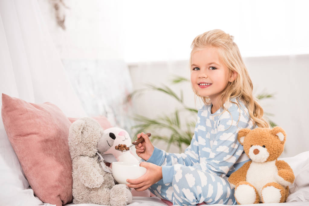 усміхнена дитина в піжамі, що годує м'які іграшки в ліжку
 - Фото, зображення